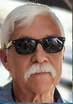 Harry M. Pritikin, an older man with white hair, a full white mustache, and dark sunglasses, outdoors in a blue collared shirt.